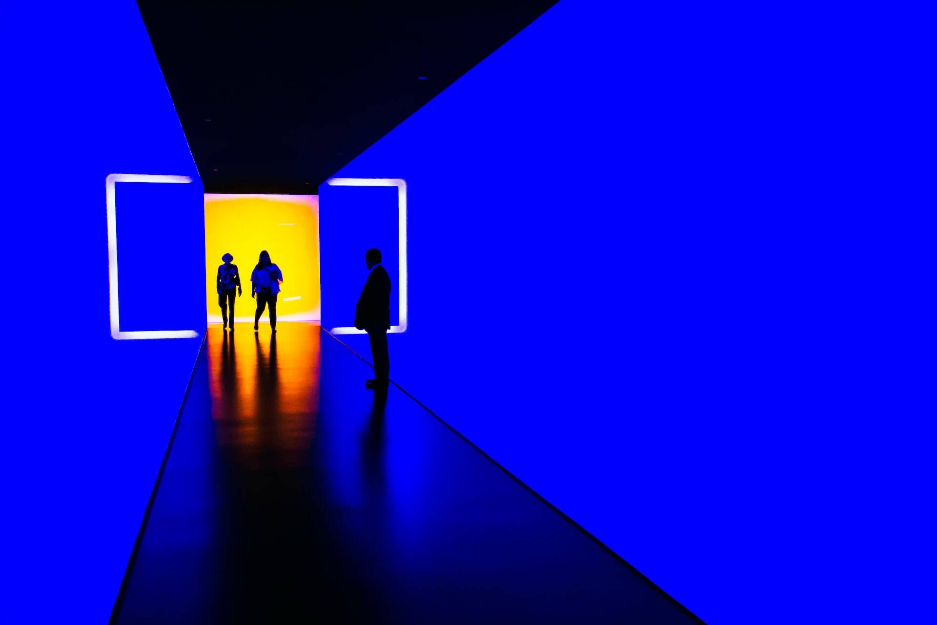 Two people walking in a blue corridor with a yellow wall at the beginning